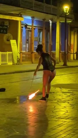 A woman wearing a black leotard and sneakers is performing with fire on a cobblestone street at night. She is holding a rope connected to a flaming object, which she is swinging. The fire is creating a large bright flame on the ground in front of her. The street is lit by a streetlamp and the warm glow of the fire and building lights. In the background, there are buildings with wooden balconies and columns. The woman is looking towards the camera and smiling.