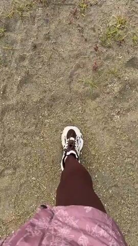 A first-person perspective video shows a person walking on a sandy surface. The person is wearing dark leggings and white and brown sneakers. A portion of a pink patterned jacket is visible at the bottom of the frame. The ground is a mix of sand and small pebbles, with patches of dry grass and some twigs scattered around. The camera angle is looking down at the person's feet and the ground as they walk.