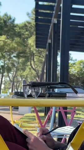 The video shows a close-up of a person's hands as they write in a notebook with a pen. They are seated at a yellow table outdoors, with other colorful chairs visible in the background. On the table, a pair of glasses, a black case, and a book with the text "MILAN" visible on its spine are placed. The scene is set amidst lush green trees, suggesting a natural, possibly park-like or coastal environment. The bright sunlight indicates it is daytime, likely a warm and pleasant day. The focus on the hands and writing implies an activity of journaling, studying, or creative work. The overall mood appears calm and focused. The setting in Ochamchire, Abkhazia, further contextualizes the scene as being in a region known for its natural beauty.