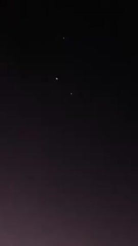 The video shows fireworks at night. There are bright bursts of light in various colors. One burst is green and another is red. There is also what appears to be a shooting star. There are a few blurry trees in the background. The sky is dark.