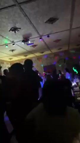 A video shot in a dimly lit room with colored party lights shows a gathering of people, mostly in silhouette. The room's ceiling is tiled with square panels and decorated with what appear to be recessed lights and decorative elements. The party lights cast shifting colors across the room, and people are standing, some moving slightly, within the space. A bright neon sign is visible. The overall impression is of a party or celebration in a private venue.
