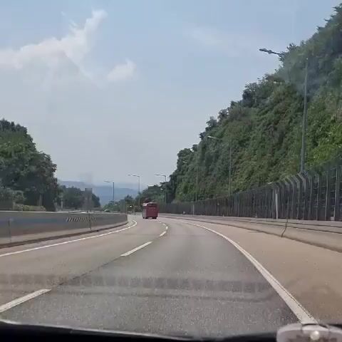 The video shows a view from the dashboard of a car driving on a highway. The highway is paved and has multiple lanes separated by white lines. On the left side of the highway, there is a concrete barrier and trees. On the right side, there is a steep, tree-covered hillside and a fence. The sky is blue with some white clouds. In the distance, on the highway, a red bus is visible. The overall environment appears to be a sunny day in a rural or mountainous area.