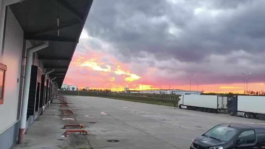 A parking lot with multiple trucks and a black van parked under a cloudy sky with a sunset in the background. The trucks are parked in a line on the right side of the frame. The van is parked on the left side of the frame. The sky is filled with dramatic clouds with streaks of orange and pink from the sunset. The parking lot is paved and appears to be wet in some areas. There is a building on the left side of the frame with a row of loading docks. The environment appears to be industrial or commercial.