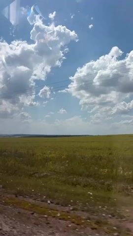 The video shows a landscape view from a moving vehicle. The sky is bright blue with large, fluffy white clouds. In the foreground, there is a field of green and yellow grass, possibly crops or wildflowers. A utility pole with wires is visible on the left side of the frame, and a circular white road sign with a black symbol is attached to it. The sign appears to have a black vertical line and two horizontal lines, possibly indicating a specific type of restriction or information. The ground in the immediate foreground, seen through the vehicle's window, is brown and appears to be a dirt or gravel road. The overall impression is a sunny day in a rural or open area.