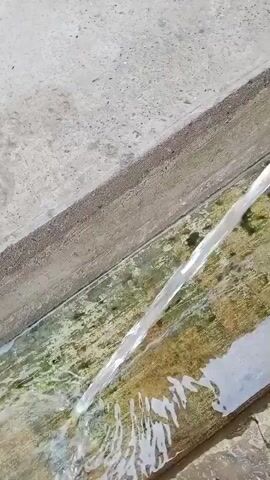 The media file shows a close-up, high-angle shot of a stream of water flowing from a white pipe into a narrow concrete channel. The channel appears to be outdoors, with a rough, textured concrete surface surrounding it. The water itself is clear and creates ripples as it enters the channel, which has some green algae or moss growing on its sides, suggesting it may be a drainage or irrigation channel. The lighting indicates it is daytime, likely sunny, given the bright illumination and slight shadows. There are no people visible in the frame. The activity is the continuous flow of water, possibly for practical purposes like irrigation or draining. The setting is man-made with concrete structures, but the presence of natural elements like water and algae suggests an environment where nature and human construction coexist. No text is visible in the media file. The overall impression is one of simple, functional movement of water in an outdoor, possibly rural or semi-urban, setting. The location context provided is Suzak, Kyrgyzstan.