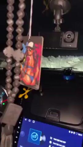 The video shows the interior of a car at night. Hanging from the rearview mirror is a novelty air freshener card featuring a woman in a blue bikini, a white hat, and sunglasses, leaning back. The card has the text “Young Legend.” A camera is mounted above the mirror, and a small, blue toy helicopter sits on the dashboard. The car's touchscreen display shows music playing.  A hand briefly moves the air freshener for a better view.