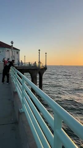 A vertical video opens on a pier railing at eye level. The railing is light blue with white accents and runs diagonally across the lower right of the frame. Beyond the railing, the ocean is a deep blue with whitecaps. In the middle ground, a pier juts out into the ocean, supported by thick concrete pillars. Several people are visible on the pier, some fishing, and one couple stands close together, leaning over the railing. The sky is a gradient of pale blue at the top fading to a warm orange and yellow near the horizon, indicating a sunset. A bright, glowing sun is visible on the right side of the frame, low on the horizon, casting a shimmering path of light across the water. A ship is faintly visible in the distance on the left side of the horizon. The camera pans slightly to the right, revealing more of the sun and the ocean.