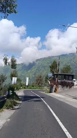 The video captures a slightly elevated point-of-view, looking down a paved road that curves to the right. The road has a white solid line on the right edge and a dashed white line in the center. On the left side of the road, there is a concrete barrier and some vegetation, including trees. On the right side of the road, there is a low stone wall, a building with a dark roof and windows, and some plants. Above the road, there are signs, including road signs indicating a curve to the right and a sign with the word "Gaia" on it. The background consists of a lush green mountainside with trees and patches of clouds obscuring the upper slopes. The sky is a vibrant blue with scattered white clouds. The lighting suggests it is daytime, likely morning or afternoon. There are no people visible in the video.