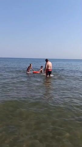 The video shows a bright, clear day at the beach. The sky is a pale blue, and the ocean water is calm with gentle waves. In the water, there are three people. One is an adult male wearing black swim trunks. He is standing in the water and facing to the left. Two children are also in the water. One child, wearing a life vest, is on the left, seemingly moving or playing in the water. The other child is closer to the adult male, also in the water. The adult appears to be interacting with the children. The water is shallow enough for the adult to be standing comfortably. The scene conveys a sense of leisure and family fun.