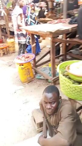 The video shows a bustling outdoor market scene. Several people are visible, some moving around and others seated. One man is sitting on the ground wearing a brown shirt and headphones. Baskets of vegetables like cabbages and green beans are clustered nearby. There are also wooden tables and other items scattered around, typical of a market environment. The general atmosphere is busy with multiple individuals engaged in what appears to be commercial activity.