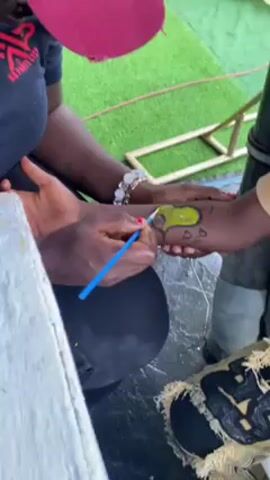 Here's a forensic description of the video:

The video depicts an outdoor event, possibly a child's birthday party, with various activities and decorations. A woman wearing a maroon cap and a black shirt with the logo R2B PARKS CLUB is seated at a white table. She is painting a design on the forearm of a young child with dark skin. The design appears to be a cartoon character, possibly from the popular cartoon "SpongeBob." Several bottles of paint, brushes, and painting supplies are on the table.  A large inflatable cartoon character is also visible in the background. The ground is covered in artificial turf. Other people are seen in the background, indicating a social gathering. The video is recorded in a close-up shot, focusing on the painting activity.