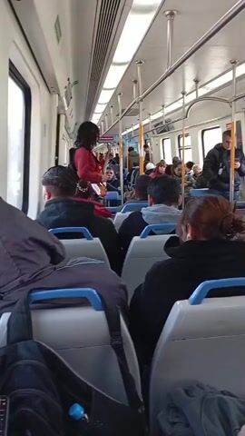 This media file shows the interior of a train car with several passengers seated and standing. The scene is brightly lit, suggesting it is daytime, and the windows reveal a blurry exterior that does not offer specific location cues. The passengers are dressed in casual attire, with some wearing jackets, indicating a cool or moderate temperature.

A woman with long dark hair, wearing a red top and backpack, stands near the center of the car, playing a guitar. She appears to be entertaining the passengers, some of whom are looking in her direction. Her focus is on her instrument, and her posture suggests she is actively engaged in playing.

The train car itself is typical of public transportation, with rows of seats in light colors, blue headrests, and overhead handrails. Bags and backpacks are visible on seats and the floor, suggesting passengers are traveling. The overall mood appears relaxed and ordinary, with people going about their commute. There is no visible text in the media file.