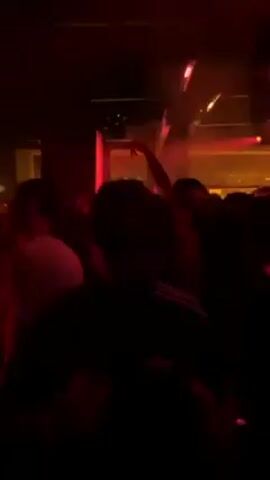 The video takes place in a dimly lit club with a red hue. A crowd of people is present, their silhouettes mostly obscured by the darkness. One individual stands out as the camera approaches them. This person is wearing sunglasses and a black jacket with white stripes. They are holding a clear plastic cup filled with liquid and a small white object, possibly a cigarette. The person is looking at the camera and appears to be talking. The background is indistinct, but there are other people moving around and some lighting fixtures visible. The overall atmosphere is lively and energetic, suggesting a social gathering or party.