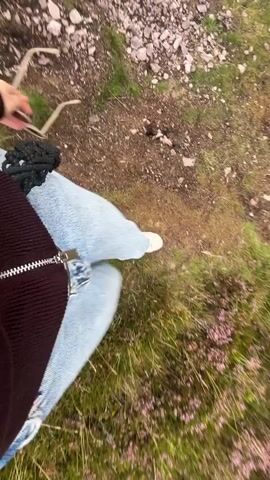 A first-person perspective video shows someone walking along a grassy, rocky path. The person is wearing light blue jeans and white sneakers. They are holding a pair of sunglasses in their right hand, dangling them by the arm. The left hand is not visible. The ground is covered with patches of green grass, brown dirt, and grey rocks, with occasional clusters of small, purple flowers. The camera angle is low, focusing on the person's lower body and the ground. The movement suggests a casual walk.