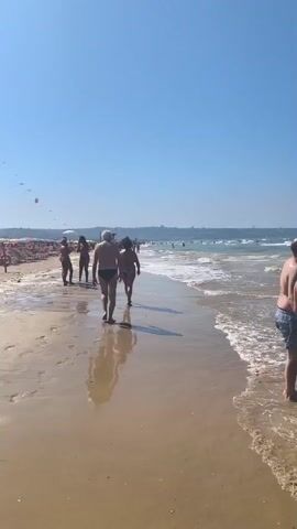 The video shows a sunny day at the beach. Several people are walking along the shoreline, with the ocean on their right and beach umbrellas and chairs on their left. The sky is clear and blue. The waves are gentle. A few people are visible further down the beach, and there are more umbrellas and chairs in the distance. There is an inflatable mattress on the sand.