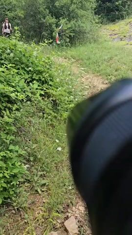 The video shows a person in the foreground holding a camera lens, obscuring a portion of the frame. In the background, a dirt path winds through a wooded area with lush green foliage. Two individuals are visible on the path, dressed in athletic wear suitable for outdoor activity. The person on the left is wearing a dark tank top and shorts, with a backpack. The person on the right is wearing a light-colored tank top and dark leggings, also with a backpack, and a hat. There is a marker with orange and white tape visible further up the path to the right, suggesting a trail or race course. The individuals appear to be moving along the path, with the person on the left running and the person on the right walking or jogging. The lighting suggests it is daytime with sunlight filtering through the trees.
