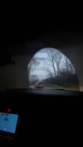 The video shows a view from inside a car driving on a narrow, paved road. Another yellow or orange car is ahead in the distance. There are leafless trees along the roadside, and the sky is overcast with a hint of light. The car's dashboard is visible, including the navigation screen and some red lights. The atmosphere appears to be somewhat gloomy.