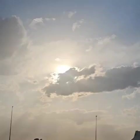The video shows a sky with the sun shining through clouds. The sun is partially obscured by a dark cloud, creating crepuscular rays that extend outwards. There are also lighter clouds scattered across the sky. In the lower part of the frame, two tall, thin poles with lights on top are visible, one on the left and one on the right. The bottom edge of the frame shows a dark, silhouetted shape that could be a building or terrain. The overall impression is of a bright but partly cloudy day.