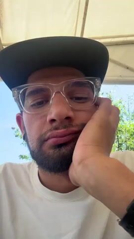 A man with a beard and glasses is in the foreground. He is wearing a white t-shirt and a dark baseball cap. He is resting his head on his hand and his mouth is open. He sticks his tongue out at the camera. The background is blurred but appears to be outdoors.