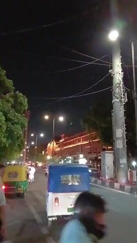 The video shows a city street scene at night. The street is illuminated by streetlights. Several vehicles, including a white and blue auto-rickshaw and a red SUV, are visible on the road. Pedestrians are walking on the crosswalk.  A large building with illuminated windows is in the background. Power lines crisscross overhead.