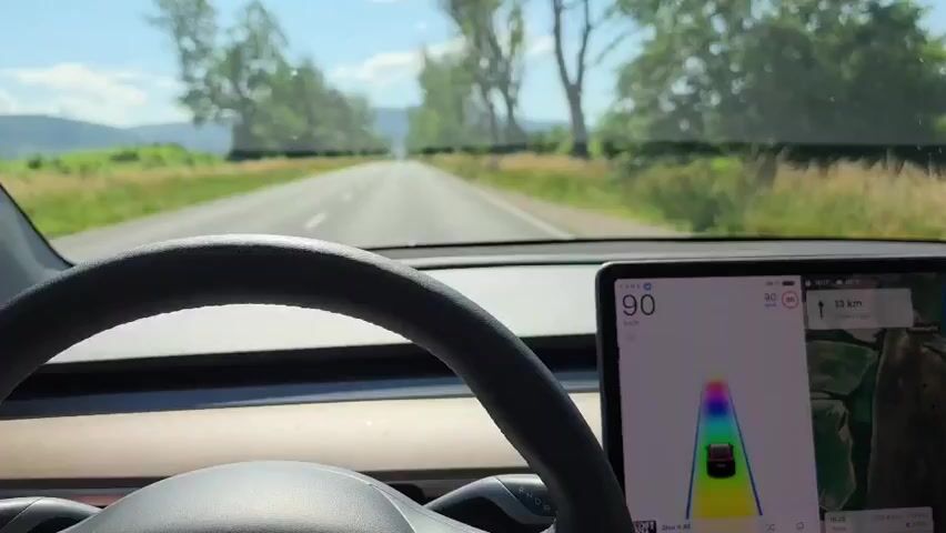The video shows a first-person perspective from inside a car, looking forward through the windshield. A black steering wheel is in the lower-left foreground. On the right, a large touchscreen display shows a speed of 90, a map, and a visualization of a car driving with a rainbow-colored path, indicating an autonomous driving mode. The car is on a straight, paved road with white lane markings. Green fields and numerous trees line both sides of the road, and distant mountains are visible. The sky is blue with scattered white clouds, suggesting a clear, sunny day. No people are visible.