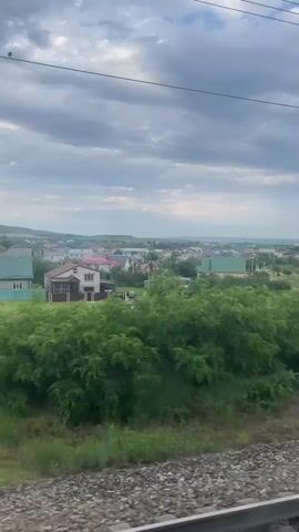 This video shows a view from a moving train traveling through the village of Zmeyka, Russia. The scene is dominated by lush green vegetation in the foreground and middle ground, with a collection of houses and buildings spread across a gentle slope in the background. The houses feature a variety of roof colors, including pink, green, and yellow, suggesting a residential area. A winding dirt road can be seen cutting through the grassy fields.

In the distance, a large, imposing mountain range rises against a cloudy sky. The clouds are heavy and gray, indicating overcast weather, perhaps with a chance of rain. The time of day appears to be late afternoon or early evening, given the diffused light and the overall subdued atmosphere. The video captures the essence of rural or suburban life in Russia, with a focus on the natural landscape and the juxtaposition of human settlement against the grandeur of the mountains.

The activity depicted is the train's journey, with the camera positioned to capture the passing scenery. The perspective from the train window creates a sense of movement and observation. There are no people or specific objects of focus other than the landscape itself. The overall mood is one of peaceful travel and scenic beauty, with a hint of dramatic grandeur from the mountain backdrop. No text is visible in the video.