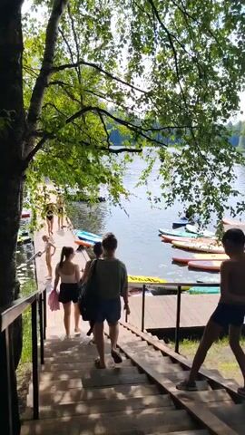 The video opens with a shot looking down a wooden staircase towards a lake. There are several people walking down the stairs. At the bottom of the stairs, to the left, is a wooden dock with several paddleboards and kayaks. To the right of the dock, there are people sitting on the grass. The video then pans to the right, showing more of the lake and people sitting on the grass. The camera then pans back to the left, showing the dock and people walking on it. The video ends with a shot of the lake and trees.