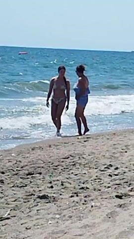 The video shows two women walking on a sandy beach near the water. The woman on the left is wearing a bikini and the woman on the right is wearing a blue cover-up. The ocean is blue and there are some buoys in the water. The sky is clear and the sun is shining.