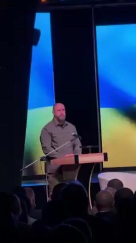 A man with a beard is standing behind a wooden podium, speaking to an audience. He is wearing a gray button-down shirt and khaki pants. The audience is sitting in front of him, and the backs of their heads are visible. Behind the man is a large screen displaying the Ukrainian flag. The room is dimly lit.