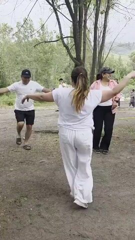 A group of four people are gathered outdoors in a wooded area with dirt ground and sparse trees. The person in the foreground, on the right, is a woman wearing white clothing. She is facing away from the camera with her arms outstretched to her sides, bent at the elbows, with her hands in fists. To her left, a man in a white t-shirt and black shorts is also facing her with his arms outstretched to his sides. He is wearing a dark baseball cap and sandals. Further to the right, a woman in a pink jacket over a white top and black pants is smiling and looking at the people in front of her. In the background, another person is visible, partially obscured by trees. The terrain slopes upwards behind the group. The lighting suggests it is daytime.
