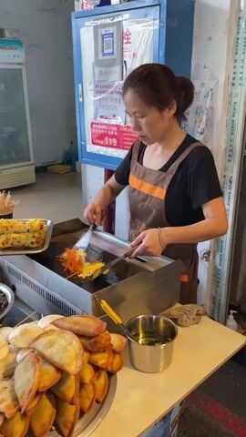 A woman wearing a brown apron is cooking food on a flat grill. She is using two spatulas to flip and move the food around. To her left, there is a tray with some kind of egg roll, a bowl with a dark liquid and some food sticking out, and a plate of lettuce. In front of her, there is a large plate piled high with fried dumplings, a plate of what appear to be steamed buns, and a metal container of oil with a brush. Behind her is a blue and white refrigerator with a sign that says air conditioning is available for free. In the background, we can see another white refrigerator and someone standing on the left side of the frame. The woman seems focused on her task.
