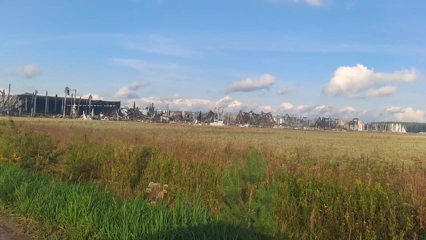 This media file captures a panoramic view of a large industrial building that has been severely damaged by fire. The structure, appearing to be a warehouse or factory, is in a state of ruin, with much of its roof and walls collapsed or missing. Charred remains of metal beams and debris are visible throughout the site, indicating the intensity of the fire. The building is situated in a rural or semi-rural setting, with a large, open field of dry grass and some vegetation in the foreground. The field extends to the base of the damaged structure. In the distance, more of the ruined building can be seen stretching across the landscape. The sky above is blue with scattered white clouds, suggesting fair weather. The time of day appears to be either late afternoon or early evening, judging by the light. The scene evokes a sense of devastation and destruction. There is no visible text within the media file. The location is contextually provided as Sluchainyi, Russia.
