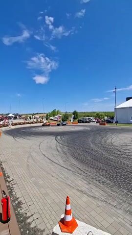 The video shows a bright, sunny day with a blue sky and scattered white clouds. The scene is an outdoor asphalt or paved area that appears to be a race track or drift course. Orange traffic cones are strategically placed around the perimeter of the course. Several vehicles are present. In the foreground, a red fire extinguisher is mounted on a brown wooden barrier. In the middle ground, a sports car, primarily orange with some black accents, is performing a drift, leaving tire marks on the ground. Behind it, another car, dark in color, is also drifting. A group of people are visible in the background, some standing near tents or structures, and others potentially spectating. There are also other vehicles parked in the distance. The overall impression is of a motorsport event in progress.