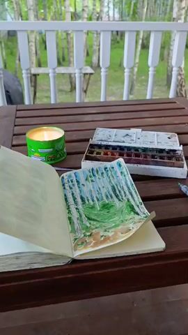 The video shows an open sketchbook with a watercolor painting of a forest scene. Next to the sketchbook is a tin of watercolor paints and a lit candle. The scene is set on a wooden table outdoors, with a white railing and trees visible in the background. The lighting suggests it is daytime. There are no people or animals visible in the video. The camera slowly zooms out, showing more of the surroundings.
