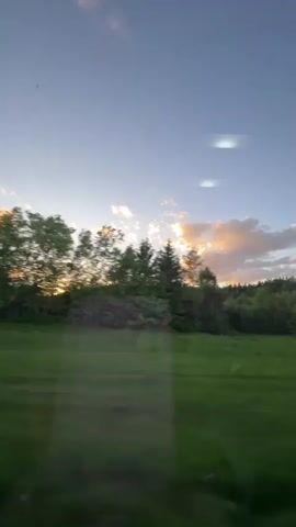 The video shows a view from a moving vehicle, traveling through a rural area. A wide expanse of green grass is visible, bordered by trees in the background. The sky is a mix of blue and orange hues, likely from a setting sun. A small, paved road is briefly visible, then the view returns to the grass and trees. The camera appears to be behind a slightly dirty window, with some reflections visible.