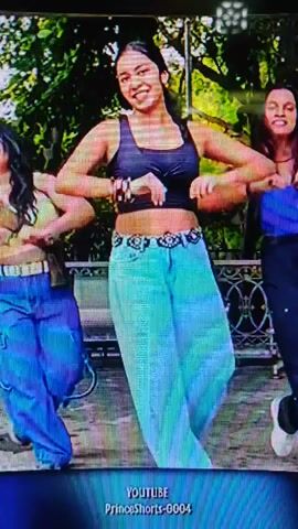 A group of three young women are dancing outdoors. The woman in the center is wearing a black sports bra and light blue baggy pants, and she is smiling and dancing energetically. The woman on the left is wearing a yellow top and blue pants, and she is also dancing. The woman on the right is wearing a blue top and black pants, and she is dancing with her mouth open. The background appears to be a park with trees and greenery. There is text overlay at the bottom of the screen that reads "YOUTUBE PrinceShorts-0004".
