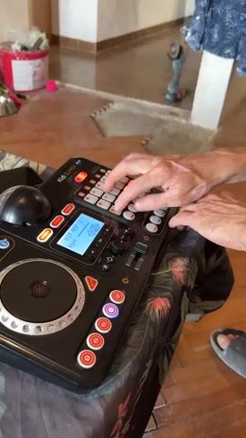 The video shows a close-up of a person's hands interacting with a black electronic musical instrument, resembling a DJ controller or a sampler. The instrument has a variety of buttons, pads, a screen, and a turntable-like disc. The person's hands are positioned over the instrument, with fingers poised to press buttons or manipulate controls. The lighting suggests an indoor environment, with a wooden floor visible in the background, along with some household items and a partially visible person in the upper right corner. The overall scene is static, with the focus solely on the interaction with the musical device.