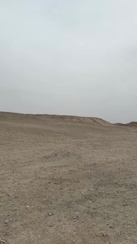 The video starts with a wide shot of a flat, gravelly desert landscape under a cloudy sky. Two figures are visible in the distance. The camera pans, showing tire tracks crisscrossing the ground, before focusing downward. The view shifts to a pair of legs in blue jeans and brown cowboy boots walking across the gravelly terrain. The edge of a black jacket is visible.