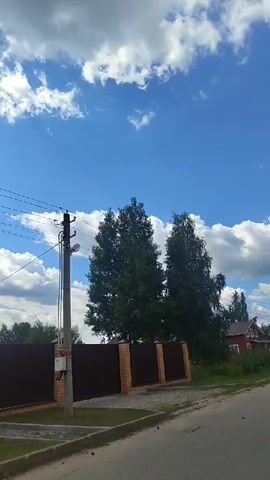 The video shows a street view with a clear blue sky dotted with fluffy white clouds. On the left, there is a tall utility pole with wires extending horizontally across the frame. A dark brown fence with brick pillars runs along the side of the road, separating it from a grassy area and trees. Further in the background, houses and trees are visible. The camera pans slowly to the right, revealing more of the houses and the street. The overall impression is a sunny day in a residential area, possibly a village or suburban setting.
