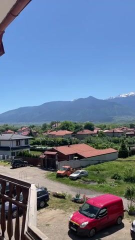 The video showcases a picturesque view of a small town nestled beneath a towering mountain range, captured on a clear, sunny day. The camera appears to be positioned on a balcony, looking over a scene composed of residential houses, vehicles, and natural landscapes. The foreground contains several vehicles parked in what seems to be a residential area, including a distinctive red van. Houses with red-tiled roofs are scattered throughout the midground, interspersed with green trees and grassy areas. In the background, a majestic mountain range dominates the horizon, with patches of snow visible on its peaks. The sky is a clear, cloudless blue, adding to the serene atmosphere of the scene. The camera slowly pans to the right, revealing a straight road lined with houses. Cars are seen moving along the road, indicating some activity in the town. Overall, the video provides a tranquil snapshot of a small town set against the backdrop of a beautiful mountain range.