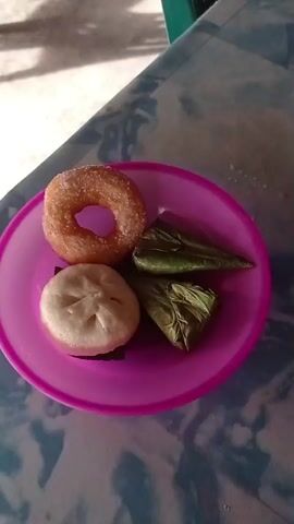 A plate containing four items of food rests on a table. From the top left and moving clockwise, the items are a ring-shaped pastry dusted with sugar, two triangular parcels wrapped in green leaves, and a round, white steamed bun. The plate is pink. The table has a patterned surface.