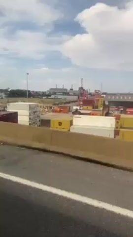 The video, shot from the passenger seat of a moving vehicle, shows a harbor scene. Stacks of shipping containers in various colors, including white, orange, yellow, and pink, line the port. In the background, a large industrial complex with smokestacks is visible. A gray vehicle passes in front of the camera vehicle. The sky is blue with scattered white clouds.  The camera is positioned alongside a concrete road barrier.  A white line marks the road.