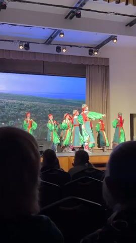 The video shows a dance performance on a stage in a room with an audience.  There are at least seven dancers dressed in green and red traditional costumes, moving in coordinated patterns. A large screen behind them displays a landscape with a blue sky, green grass, and red flowers. The audience sits in rows of chairs, observing the performance.  The room has a modern ceiling with recessed lighting.  The perspective is from within the audience.