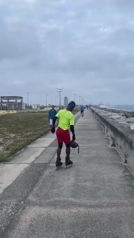 A person is rollerblading on a paved path next to a rocky embankment. The person is wearing a neon green shirt, red shorts, and a helmet. Another person is walking on the path towards the rollerblader, wearing a blue shirt and white shorts. The sky is overcast and the ocean is visible to the right. In the distance, there are buildings and streetlights.
