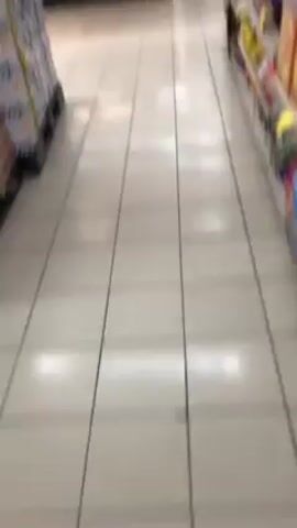 The video shows a person walking down the snack aisle in a grocery store. The camera is pointed at the shelves, which are full of various snacks, including chips, candy, and cookies. The floor is tiled and there are stacks of boxes in the background.  A person can be briefly seen in the background standing behind the snack shelves.