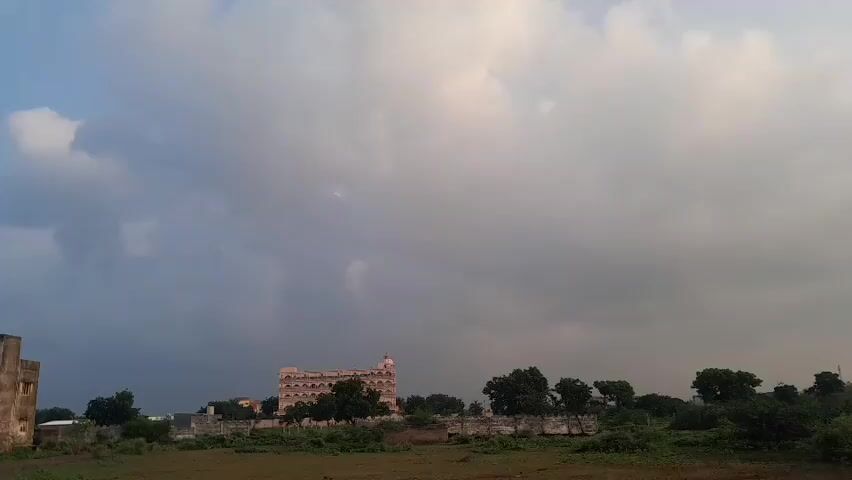 A video shows a wide shot of a rural landscape with a cloudy sky. In the foreground, there is dry, brown grass. In the middle ground, there are several buildings and trees. On the left, a two-story building is visible, with some crumbling sections and a water tank nearby. Further to the right, a larger, ornate building with multiple levels and arched windows stands out. A line of trees and a low wall separate this building from the foreground. More trees and smaller structures are scattered across the landscape. Power lines and poles are visible in the sky. The overall scene suggests a somewhat undeveloped or perhaps historical area under a dramatic, cloudy sky.