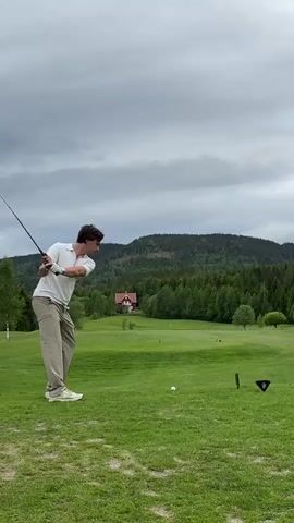 The video shows a man playing golf on a green golf course with a red house and lush green trees visible in the background under an overcast sky. The man, dressed in a white shirt and light brown pants, swings a golf club, hitting a golf ball that is situated on the tee. After the shot, he watches the ball's trajectory before resting the club on his shoulder.