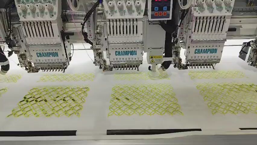 The video shows a close-up, high-angle shot of a multi-head embroidery machine in operation. The machine is large and white, with multiple embroidery heads labeled "CHAMPION." Each head has numerous needles positioned above a white fabric. The fabric is stretched taut on a frame and is in the process of being embroidered with a yellow, interlocking, lattice-like pattern. The embroidery appears to be a continuous run, with several completed sections of the pattern visible. The lighting is bright and even, highlighting the details of the machine and the embroidery. The camera slowly pans to the right, showing more of the fabric and the ongoing embroidery. There are no people visible in the video.