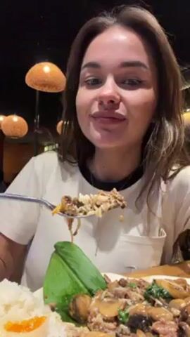 A woman is eating a plate of food. She is wearing a white t-shirt and a silver necklace. She holds a fork full of food up to her mouth and eats it. The food consists of rice, mushrooms, and some kind of meat. There is a green leaf garnish on the plate. The background is blurry, but it looks like she is in a restaurant. There are several globe-shaped lamps hanging from the ceiling. After chewing, the woman holds up her hand in a gesture that seems to indicate the food is good.