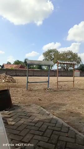 A slightly shaky, vertical video filmed from a low angle shows an outdoor scene under a bright, partly cloudy blue sky. In the foreground, a patterned brick surface is visible on the left and extends toward the bottom of the frame. To the left of the brick surface, a dark, cylindrical object, possibly a metal barrel or bin, is partially visible. The middle ground is a dry, dusty field with sparse, brown vegetation. Two open-sided, roofed structures stand in the field; the one on the left has a metal roof supported by blue metal poles, and the one on the right has a roof with visible shingles supported by wooden poles. Behind the structures, a stone or brick wall extends horizontally across the frame. Beyond the wall, trees and buildings with red tile roofs are visible. The video is tagged with "Infinix HOT 40 Pro."